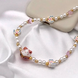 Puede incluir: Un adorno de teléfono con cuentas de perlas blancas, cuentas doradas y abalorios de colores. Los abalorios incluyen mariposas, gemas rosas y una figura de gato de la suerte. El adorno está sujeto a una funda de teléfono beige.