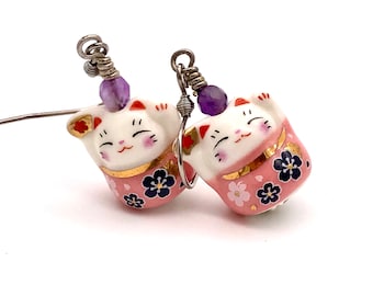 以下が含まれることがあります： 紫色の宝石のアクセントが付いたピンクと白のセラミック製の猫のイヤリングのペア。猫は伝統的な日本の幸運のお守りである、座って足を上げたポーズをしています。