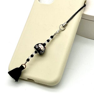 Può includere: Una custodia per telefono color crema con un ciondolo per telefono con perline bianche e nere. Il ciondolo presenta un nappa nera, una perlina a forma di gatto in ceramica e perline nere e trasparenti. Il ciondolo è attaccato alla custodia del telefono con un cordino nero.