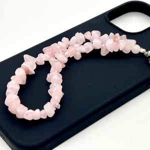 Pode incluir: Um amuleto de telefone de quartzo rosa rosa com um fecho prateado e uma alça elástica rosa. O amuleto é feito de pequenas peças irregulares de quartzo rosa.