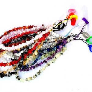 Puede incluir: Una colección de hebras de cuentas de chips de piedras preciosas de varios colores, incluyendo rojo, rosa, blanco, negro, amarillo, azul y morado. Las hebras están atadas juntas con una cadena plateada y anillos de silicona de colores.