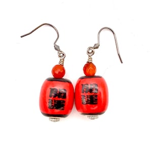May include: A pair of red dangle earrings with black Chinese characters and silver accents. The earrings are made of wood and have a red carnelian bead at the top.