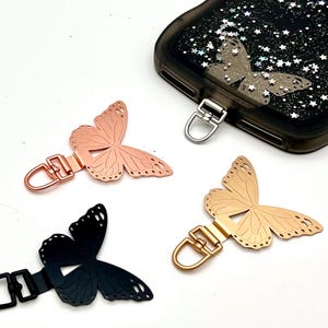 Puede incluir: Abalorios para teléfono con forma de mariposa en oro rosa, negro y dorado. Cada abalorio tiene un cierre metálico. También se ve una funda de teléfono negra con un diseño de mariposa y purpurina.