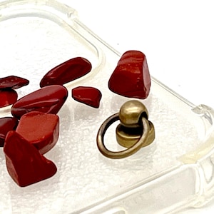 May include: Close-up of a clear phone case with red, polished stones and a brass-colored ring attachment. The stones vary in size and shape, with a glossy finish. The ring attachment has a circular design.