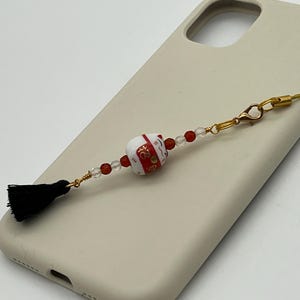May include: A white and red ceramic charm with gold accents, featuring a black tassel and red and clear beads. The charm is attached to a gold phone strap and rests on a beige phone case.