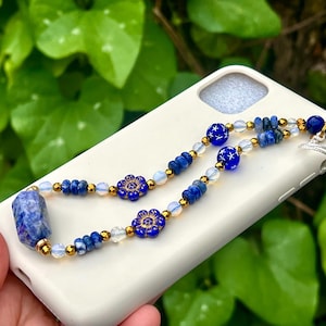 May include: A white phone case with a blue and gold beaded phone charm. The charm features blue flower beads, blue star beads, and a large blue stone. The beads are strung together with gold-colored accents.