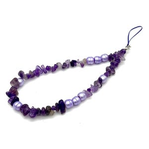 Puede incluir: Un charm de teléfono hecho a mano con chips de amatista y perlas de color lavanda. El charm tiene un lazo azul oscuro para sujetarlo. Las piedras preciosas y las perlas están ensartadas, creando un accesorio único.