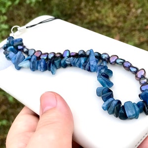 Puede incluir: Un charm para teléfono con una doble hilera de cuentas de cianita azul y perlas negras. El charm está unido a una funda de teléfono blanca.