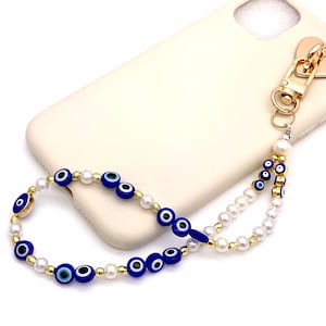 Puede incluir: Un charm de teléfono dorado con un cierre en forma de corazón y una cadena de cuentas con perlas blancas y cuentas azules de ojo turco.