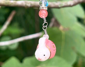 Könnte beinhalten: Ein handgefertigter Anhänger mit einem rosa-weißen Hasenanhänger mit goldenen Akzenten, der an einer silbernen Kette mit rosa und klaren Perlen befestigt ist. Der Anhänger ist für die Schmuckherstellung oder als dekoratives Accessoire gedacht.
