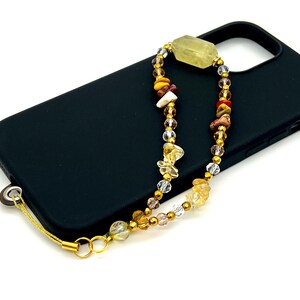 May include: A black phone case with a gold chain and beaded phone strap. The strap features a variety of colorful beads, including clear, gold, and brown.