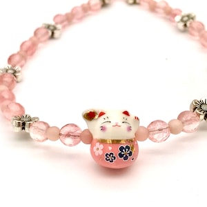 May include: A pink bracelet with a white ceramic cat charm. The bracelet is made of pink beads and silver flower charms.