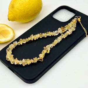 Puede incluir: Una funda de teléfono negra con un dije de teléfono de piedras preciosas amarillas. El dije está hecho de pequeñas piedras amarillas irregulares ensartadas. Un limón entero y una rodaja de limón están al lado del teléfono.