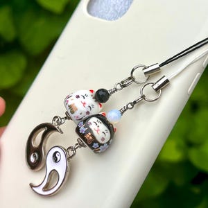 May include: A phone charm featuring two ceramic maneki-neko cat beads, one white and one black, paired with two yin-yang metal charms. The charms are attached to black and white cords with silver-toned metal rings and small accent beads.