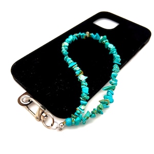 May include: A black phone case with a turquoise stone bead phone strap. The strap is made of small, irregular turquoise stones strung together. A silver-colored clasp attaches the strap to the phone case.