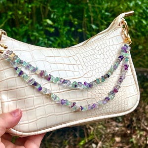Puede incluir: Bolso de mano color crema con textura de cocodrilo, herrajes dorados y una cadena decorativa hecha de pequeñas piedras preciosas de colores. Las piedras preciosas son de color púrpura, verde y transparente.