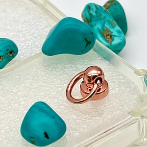 Puede incluir: Primer plano de una funda de teléfono transparente con un anillo magnético de oro rosa y varias piedras turquesas. Las piedras tienen marcas marrones y negras. El anillo está unido a una pequeña base redonda.