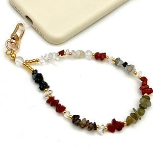 May include: A phone charm with a gold-coloured clasp and fittings. The charm features a strand of colourful, irregularly shaped gemstones, including red, black, clear, and green stones, interspersed with small gold beads.