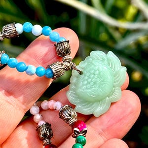 May include: A multi-colored beaded bracelet with a green jade flower charm. The bracelet features beads in shades of blue, pink, red, and green. The jade flower charm is carved with intricate details.