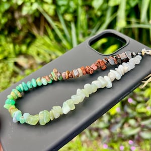 May include: A multi-colored beaded phone charm featuring green, brown, red, and white stones. The charm is attached to a black phone case. The background is blurred green foliage.