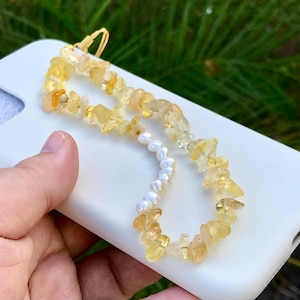 Puede incluir: Una funda de teléfono blanca con una correa de teléfono de cuentas amarillas y blancas. La correa está hecha de pequeñas piedras de forma irregular y perlas blancas.