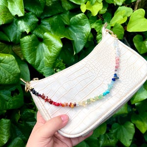 Puede incluir: Un bolso blanco texturizado con una correa de piedras preciosas de colores. La correa presenta un degradado de colores, incluyendo rojo, naranja, amarillo, verde, azul y morado. El bolso tiene herrajes dorados y un patrón de piel de cocodrilo.