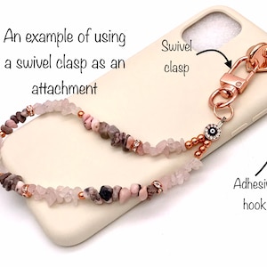 Puede incluir: Primer plano de una funda de teléfono con una correa de teléfono de cuentas rosadas y marrones unida. La correa tiene un cierre giratorio y un amuleto de ojo turco. El texto "An example of using a swivel clasp as an attachment" y "Swivel clasp" y "Adhesive hook" es visible en la imagen.