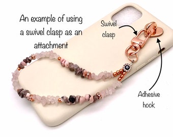 May include: A close-up of a phone case with a pink and brown beaded phone strap attached. The strap has a swivel clasp and an evil eye charm. The text "An example of using a swivel clasp as an attachment" and "Swivel clasp" and "Adhesive hook" are visible in the image.