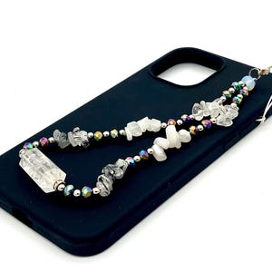 May include: A black phone case with a crystal and bead phone strap. The strap is made of white and clear crystals, iridescent beads, and silver metal beads.