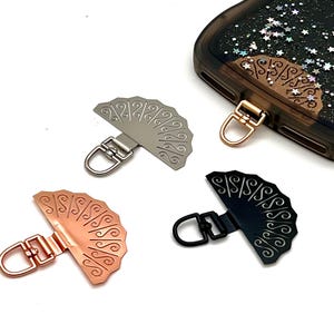 May include: Decorative fan-shaped phone charms in silver, rose gold, and black, with a matching charm attached to a phone case. The charms have a decorative pattern and a metal loop for attachment. The phone case is clear with a glittery design.
