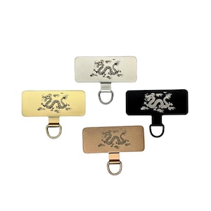May include: Four rectangular phone grip holders in gold, silver, bronze, and black. Each features a detailed dragon design and a D-ring for attachment. The grips are displayed against a white background.