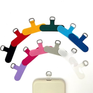 May include: A collection of colorful phone charms in various hues, including red, yellow, green, blue, pink, gray, purple, and white. Each charm features a loop for attachment and a metal ring. A phone is visible at the bottom.