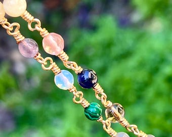 May include: Close-up of two gold-colored beaded necklaces. The necklaces feature a variety of colorful gemstones, including pink, blue, green, and clear beads. The beads are strung on gold wire, creating a delicate and elegant design.