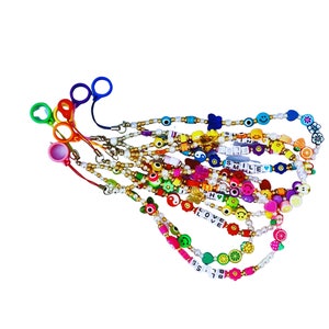 Puede incluir: Una colección de coloridas cadenas para gafas con varios dijes, incluyendo caras sonrientes, frutas y las palabras "LOVE" y "SMILE". Las cadenas presentan una variedad de colores y diseños, con un lazo al final para sujetar las gafas.