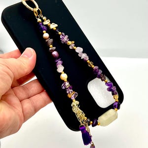 May include: A beaded phone charm with amethyst, pearl, and gold accents. The charm features a gold clasp, a purple tassel, and a large, rectangular, pale yellow stone. The charm is attached to a black phone case.