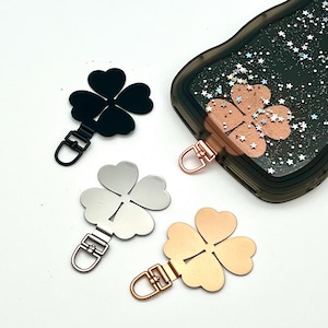 Pode incluir: Pingentes de trevo de quatro folhas em preto, prata e dourado, com uma capa de telefone a condizer. Cada pingente tem um fecho de metal para fixação. A capa do telefone é preta com um design brilhante e um detalhe de trevo.