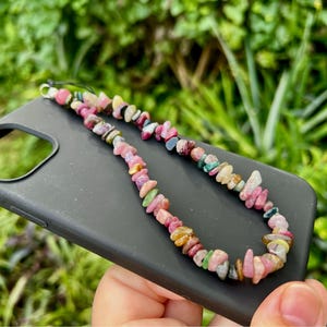 Könnte beinhalten: Eine schwarze Handyhülle mit einem bunten, perlenbesetzten Handy-Anhänger. Der Anhänger besteht aus kleinen, unregelmäßig geformten Steinen in Rosa-, Grün-, Gelb- und Weißtönen. Die Handyhülle wird in einer Hand vor einem verschwommenen grünen Hintergrund gehalten.