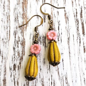 May include: A pair of drop earrings featuring a mustard yellow, ribbed, teardrop-shaped bead topped with a small, light pink, five-petaled flower bead. The components are connected by antique bronze-toned metal findings and hook ear wires.