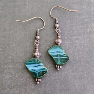 May include: A pair of dangle earrings featuring irregular, dark green glass beads with white swirling patterns. The beads are suspended from copper-toned metal hooks by a series of small, textured copper-toned metal beads and wire loops.