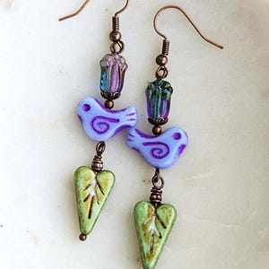 May include: A pair of handcrafted earrings featuring a bird and leaf design. The earrings have copper-colored hooks and connecting wires. Each earring has a light purple bird bead, a green leaf bead, and a flower-shaped bead with blue and purple hues.