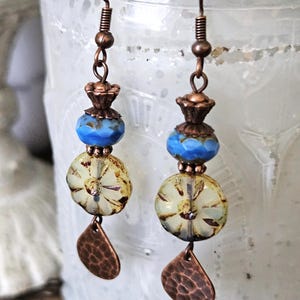 May include: Handmade earrings with copper-colored hooks and accents. Each earring features a blue bead, a yellow and white floral bead, and a textured copper teardrop charm. The earrings are designed for pierced ears.