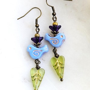 May include: A pair of dangle earrings featuring bronze-toned hooks, a dark purple flower-shaped bead, a light blue bird-shaped bead with copper-colored swirl detailing, and a translucent lime green heart-shaped bead with a gold-toned leaf design.