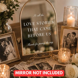 May include: An arched gold metal frame with a stand, displaying the white text: To all the LOVE STORIES THAT CAME BEFORE US Thanks for paving the way. The frame is surrounded by framed black and white photographs, candles, and white flowers.
