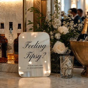 May include: A rectangular mirror sign with rounded corners featuring black text that reads Feeling Tipsy? PLEASE TAKE CARE OF OUR BARTENDERS. The sign is displayed on a marble bar next to a glass vase of white flowers and a glass jar filled with cash.