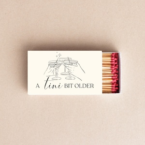 May include: A white matchbox with a black line drawing of four hands holding up glasses of champagne. The text "A Tini Bit Older" is printed on the box.
