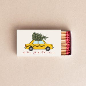 May include: Matchbox with a yellow taxi carrying a Christmas tree on top. The matchbox has the text "A New York Christmas" in red script. The matches have red tips.