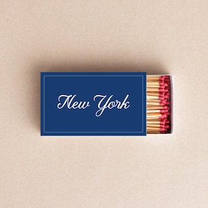 Puede incluir: Una caja de cerillas azul marino con la inscripción "New York" en escritura blanca. La caja está abierta, revelando cerillas de madera con puntas rojas.