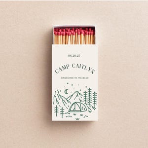 May include: A white matchbox with a green illustration of a mountain range, a tent, and stars. The text "Camp Caitlyn" and "06.20.25 Bachelorette Weekend" is printed on the box.