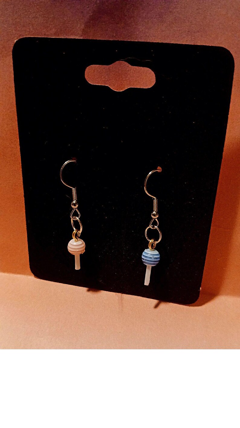 May include: A pair of silver dangle earrings with a lollipop charm. One lollipop is pink and white, the other is blue and white.