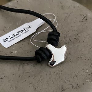 May include: A silver-colored pendant in the shape of a hammer is strung on a black cord. The pendant is the focal point, with a tag that reads "BARBARIANN history inspired jewelry" in the background.